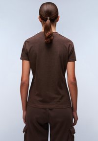 T-shirt in cotone marrone, maniche corte, design aderente, texture liscia, senza grafiche, presenta una piccola etichetta sulla spalla sinistra.