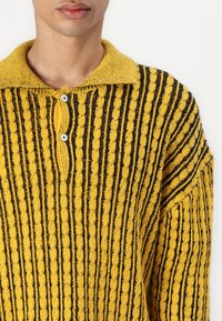 Pull en tricot jaune et noir avec une texture côtelée verticale, doté d'un col et de trois boutons au niveau du cou.