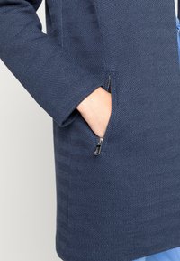 Abrigo azul marino con textura de espiga, que cuenta con un bolsillo con cremallera y mangas largas. El material parece tejido y resistente.