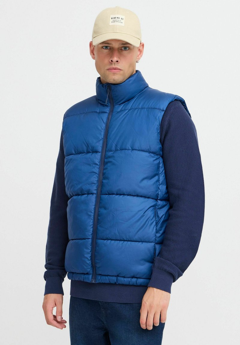 Zila polsterēta veste ar vertikālām šuvēm, augstu apkakli un rāvējslēdzēju, uzvilkta virs jūras zila teksturēta garām piedurknēm krekla.