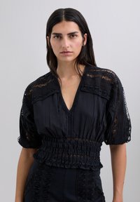 Blouse noire avec un décolleté en V profond, détails en dentelle complexes, manches bouffantes et smocks froncés à la taille pour une finition texturée.