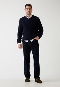 Uomo in piedi con un maglione lavorato a maglia blu navy, camicia bianca, jeans scuri e mocassini marroni su uno sfondo chiaro e semplice.