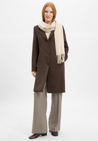 Donna che indossa un lungo cappotto marrone con bottoni, una sciarpa beige, pantaloni a quadri a gamba larga e scarpe marroni scure, in piedi di fronte a uno sfondo chiaro e semplice.