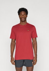 Κόκκινο κοντομάνικο μπλουζάκι με στρογγυλή λαιμόκοψη, φτιαγμένο από βαμβάκι. Διαθέτει ένα μικρό πράσινο λογότυπο στο αριστερό μέρος του στήθους. Φοριέται με σκούρες βερμούδες.