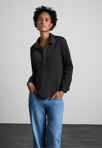 Maglietta nera a maniche lunghe con colletto, caratterizzata da una patta con bottoni e maniche arrotolate, abbinata a jeans a gamba larga azzurri chiari.