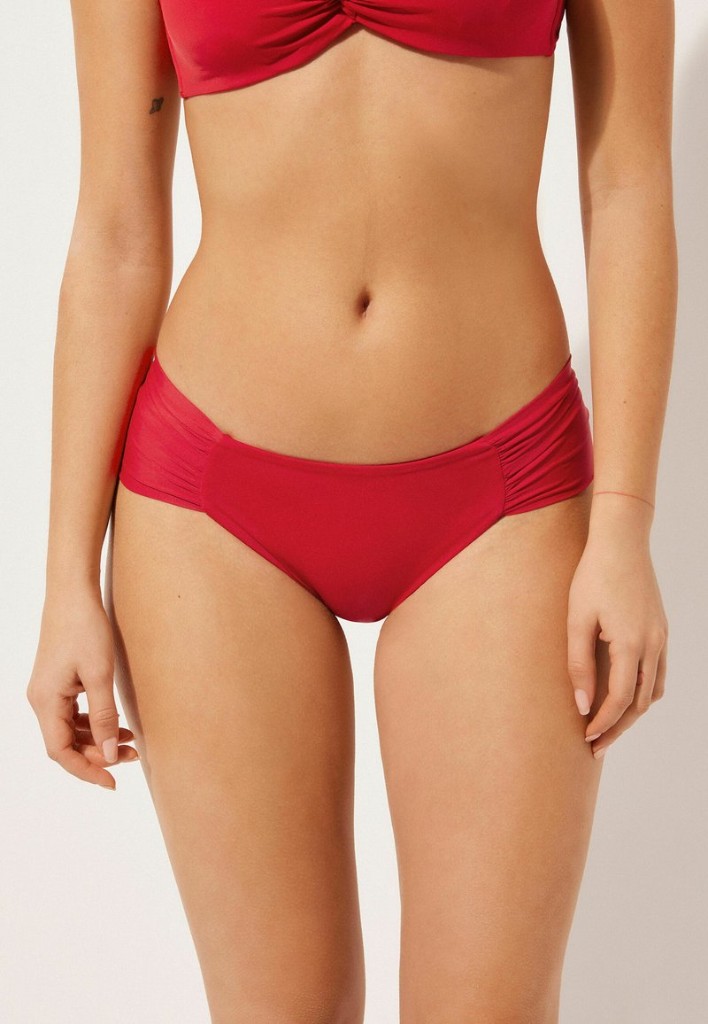 Červené bikiny s nařasenými postranními panely, vyrobené z elastického materiálu. Mají středně vysoký pas a hladký povrch.