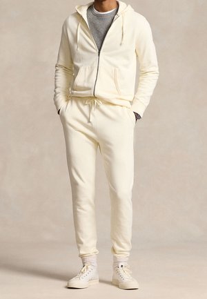 Homme portant un sweat à capuche crème zippé et un jogging assorti avec des baskets blanches montantes, debout les mains dans les poches.