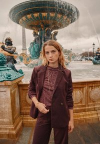 Bordeaux-kordfløyelsdrakt med en strukturert topp. Modellen står ved en utsmykket fontene, som har akvamarinfarge og gyldne detaljer, mot en skyet bakgrunn.