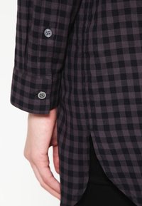 Camicia a scacchi nera e viola con polsini abbottonati, fessure laterali e una texture di tessuto liscia, che presenta un motivo classico.