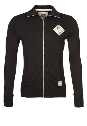 Czarna kurtka na zamek, z długimi rękawami, wysokim kołnierzem oraz beżową naszywką z logo na lewej stronie klatki piersiowej i małą metką przy dolnej krawędzi.