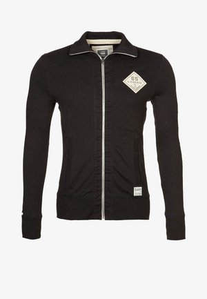 Schwarze Jacke mit Reißverschluss, langen Ärmeln, hohem Kragen und beigem Logo-Patch auf der linken Brust sowie kleinem Etikett nahe dem Saum.