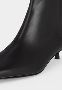 Bottine noire en cuir lisse avec un bout pointu et un petit talon bas, présentée sur un fond uni.