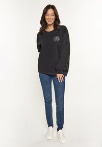 Sudadera negra con un diseño de contorno floral en el pecho, corte relajado, puños acanalados, combinada con vaqueros azules y zapatillas blancas.