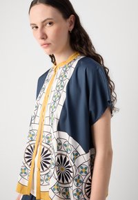 Mygtukinė blusa su tamsiai mėlyna ir geltona spalvotai geometrinėmis raštais balta, žalia ir juoda ant lengvos medžiagos.