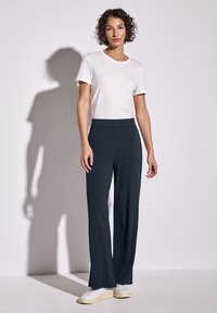 Vit t-shirt med rund hals i kombination med mörka, vida byxor. Byxorna har en slät textur och en lätt glans. Modellen står på en ljus bakgrund.