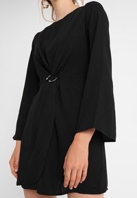 Svart klänning med en vriden frontdetalj, prydd med en silverring. Den har långa, utsvängda ärmar och ett mjukt, texturerat tyg.