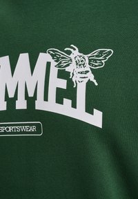 Gráfico de abeja blanca sobre texto blanco "MMEL" en tela verde oscuro, con pequeño "SPORTSWEAR" en un recuadro blanco contorneado abajo.