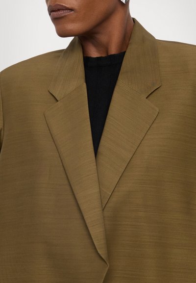 Sacou ajustat, de culoare verde olive, cu o textură netedă, revere ascuțite și o croială lejeră, purtat pe un tricou negru cu dungi. Accentuare cu cercei argintii.