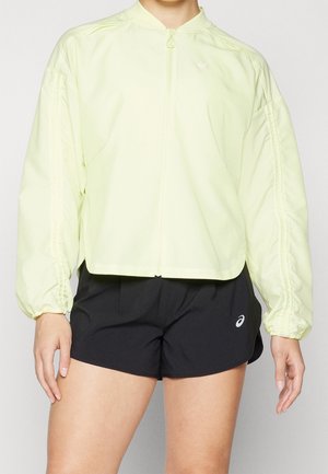 Persoon draagt een lichtgele jas met rits en elastische manchetten en zwarte sportshorts met een wit logo op de linker dij.