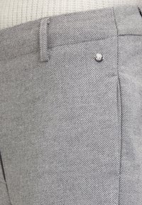 Grijze textuurbroek met een subtiele visgraatpatroon, een zijzak en een klein metalen logoaccent nabij de tailleband.