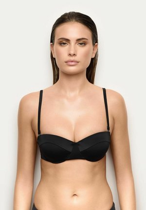 Donna con capelli bagnati che indossa un reggiseno nero con ferretto e spalline sottili regolabili, su uno sfondo chiaro uniforme.