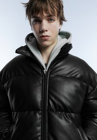 Black leather puffer jacket with a high collar and zipper closure, layered over a gray hoodie. Smooth texture and quilted pattern visible.