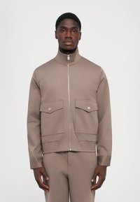 Man wearing beige zip-up jacket with two front flap pockets and matching pants, standing against a plain light background.