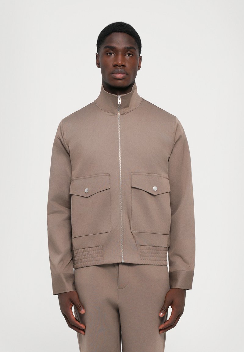 Man wearing beige zip-up jacket with two front flap pockets and matching pants, standing against a plain light background.
