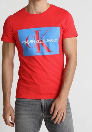 Mann trägt ein rotes T-Shirt mit kurzen Ärmeln, darauf ein blaues Rechteck und ein großes rotes "cK" sowie weißer Schriftzug "CALVIN KLEIN JEANS", kombiniert mit grauen Jeans.