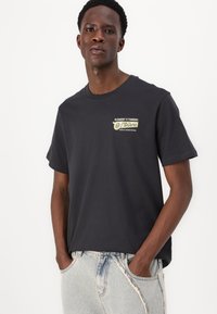 Black cotton t-shirt with short sleeves and a crew neckline. Features a printed logo in white and green on the upper left side.