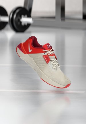 Lyse beige og rød atletiksko med snørebånd, med hvid swoosh-logo, vist svævende over en reflekterende gulv med sløret fitnessudstyr i baggrunden.