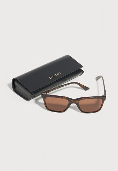 Lunettes de soleil rectangulaires teintées marron avec monture écaille de tortue à côté d'un étui fermé en cuir noir Gucci sur fond blanc.