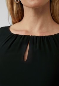 Abito nero con scollatura arricciata e dettaglio a chiave sul petto. Tessuto liscio con leggera lucentezza, silhouette aderente.