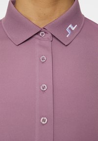 Lila polotröja med en texturerad krage, fem knappar och en liten vit logotyp på kragen. Slät tyg med en matt finish.