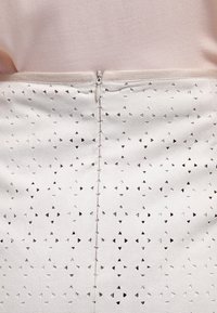 Gros plan sur une jupe blanche avec de petites découpes triangulaires et une fermeture éclair centrale, portée avec un haut rose pâle.