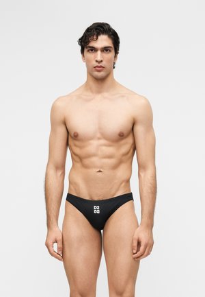 Hombre musculoso con cabello oscuro de pie contra un fondo liso, vistiendo un bañador tipo slip negro con el logo blanco "D2" en el frente.