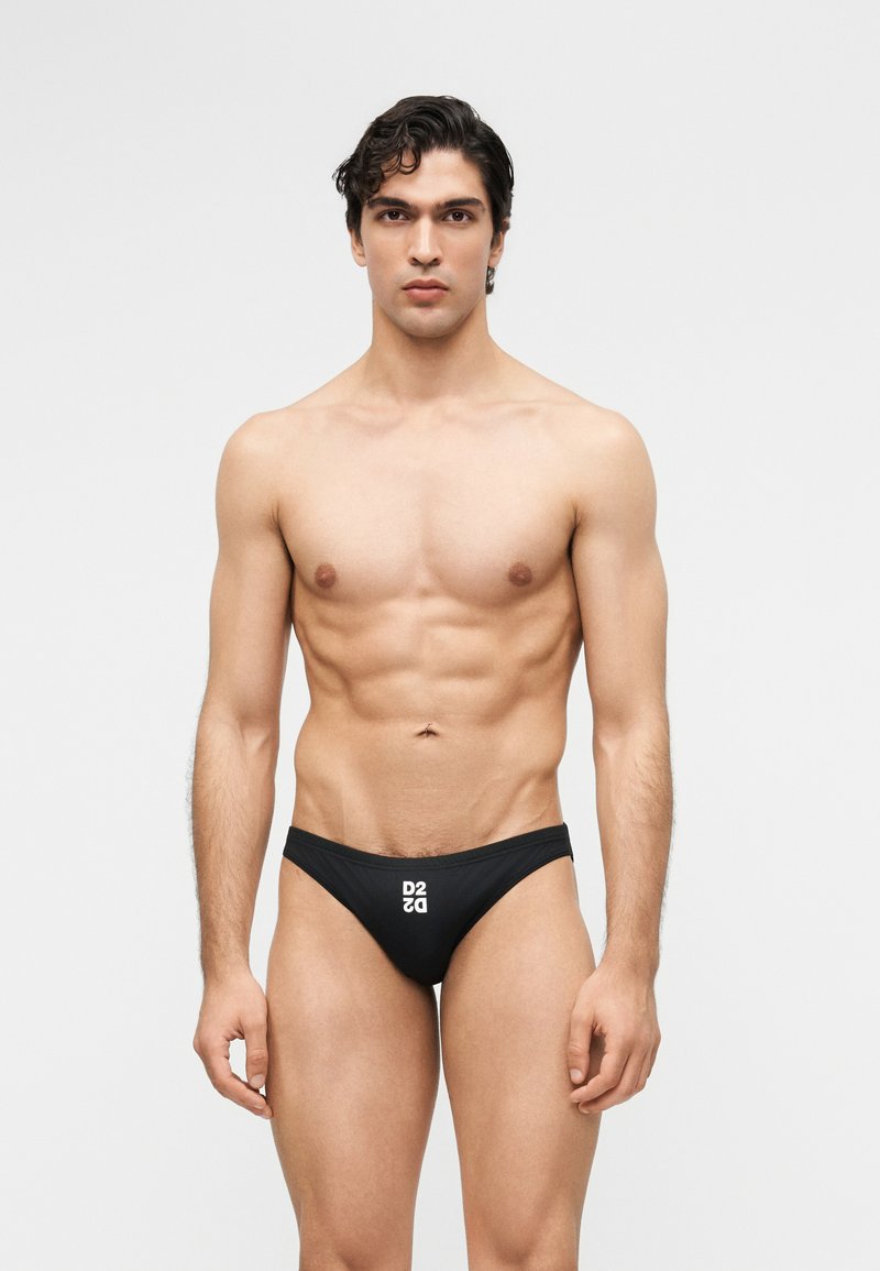 Μυώδης άνδρας με σκουρόχρωμα μαλλιά, στέκεται μπροστά σε απλό φόντο, φορώντας μαύρα μαγιό τύπου σλιπ με λευκό λογότυπο "D2" στο μπροστινό μέρος.