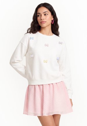 Ung kvinde iført hvid sweatshirt med små pastelfarvede sløjfer og lyserødt nederdel med prikker, står med hånden på hoften og kigger til siden.