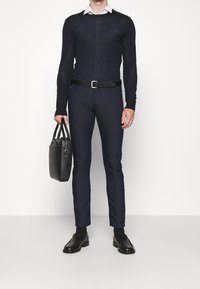 Marinblå långärmad tröja, slim-fit mörka byxor, svart bälte, svarta loafers, hållande en svart texturerad toteväska. Vit skjorta med krage under.