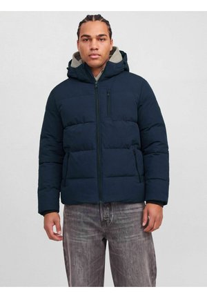 Homme portant une doudoune bleu marine à capuche et un jean gris, debout devant un fond uni gris clair.