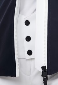 Námořnicky modrá a bílá bunda s předním zapínáním na zip, třemi černými knoflíky a hladkou texturou. Zvýrazněna kontrastními materiály.