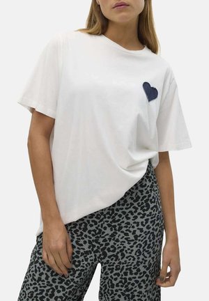 T-shirt blanc en coton à manches courtes, avec un patch en forme de cœur bleu marine sur la poitrine gauche. Porté avec un pantalon gris à imprimé léopard.
