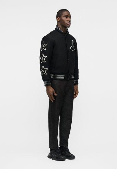 Άνδρας που φοράει μαύρο varsity μπουφάν με λευκά αστέρια και μπαλώματα με το γράμμα Β, σκοτεινό παντελόνι και μαύρα αθλητικά παπούτσια, στέκεται μπροστά σε απλό φωτεινό φόντο.