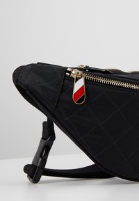 Sac banane en tissu matelassé noir avec fermeture éclair dorée et languette zippée rayée rouge, blanche et noire sur une surface blanche.