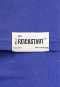 Blauwe stof met een gladde textuur. Een wit label toont "REICHSTADT™", met ontwerprichtlijnen en website in zwarte tekst.