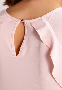 Blusa rosa chiaro con dettaglio a forma di chiave sulla schiena, bordata da morbidi volant e chiusa con un bottone dorato. Tessuto morbido.