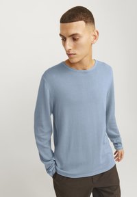 Lichtblauw langemouwen shirt van een gladde, lichte stof. Het shirt heeft een ronde halslijn en aansluitende manchetten, gedragen met donkere broeken.