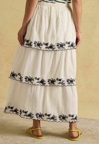 Vzdušná biela sukňa s čiernou kvetinovou výšivkou na troch vrstvených úrovniach, so zvlneným lemom a elastickým pásom.