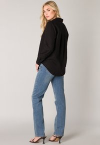 Zwarte lange mouwen shirt met een kraag, gecombineerd met lichtblauwe rechte jeans en zwarte haksandalen. Neutrale achtergrond.