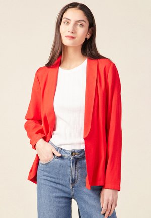 Rote, leichte Blazerjacke mit offener Vorderseite, langen Ärmeln und lockerem Schnitt, kombiniert mit einem weißen gerippten Oberteil und blauen hochgeschnittenen Jeans.